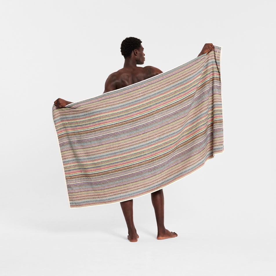 A male model standing with his back to camera, holding up a multicoloured stripe beach towel behind his back
