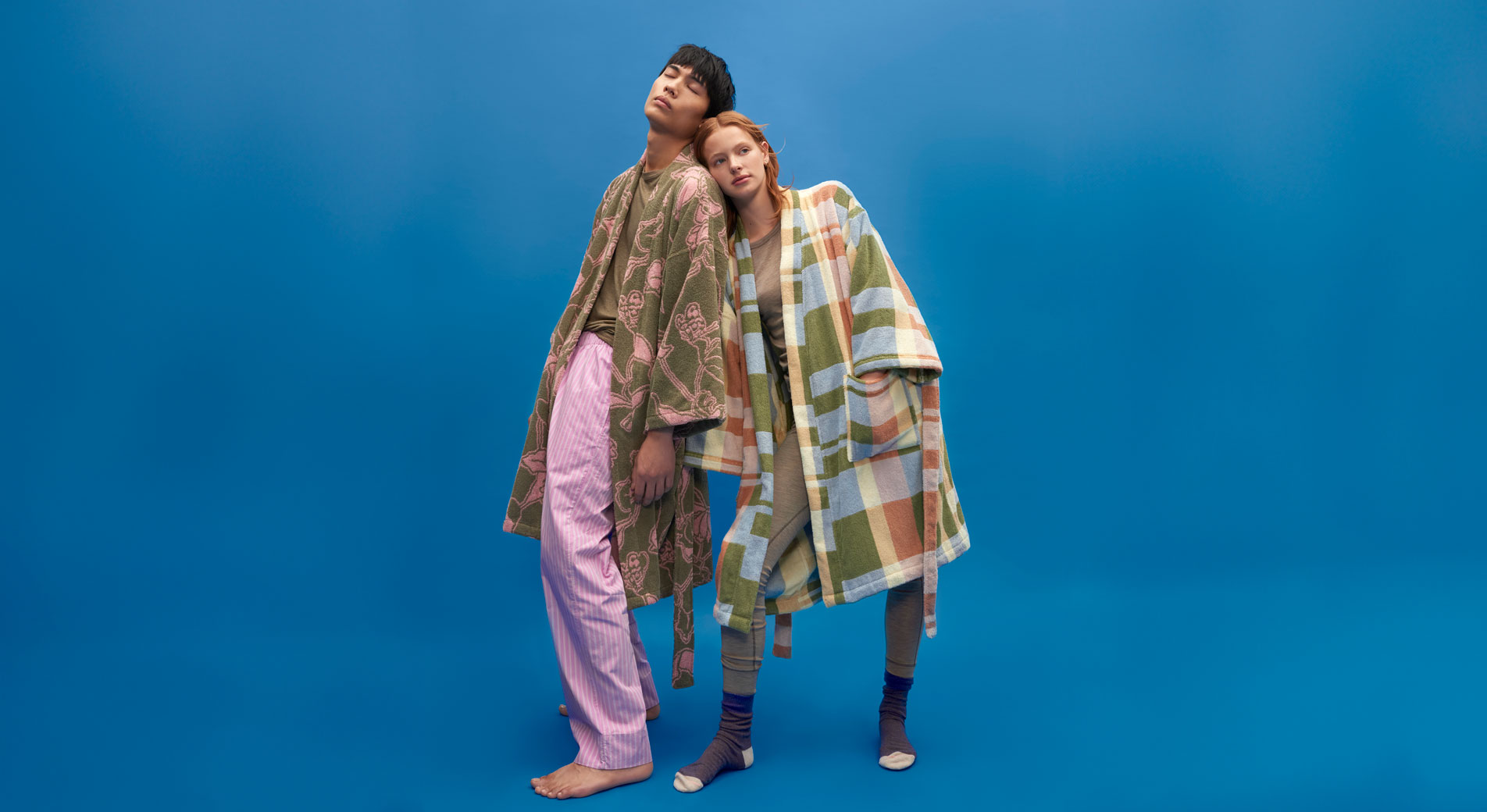 Young man and woman stand in front of a blue backdrop, both wearing stylish bathrobes. 