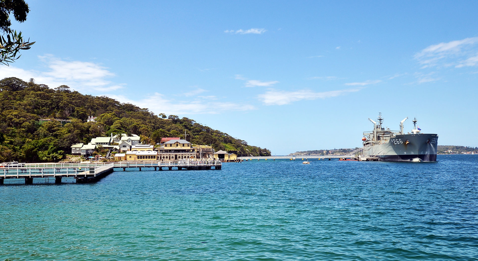 shot of chowder bay. naval ship on left side. right side, jetty, wharf, pavilion and bushland on right side. forefront of image is bright blue water. blue sky has sparse clouds.