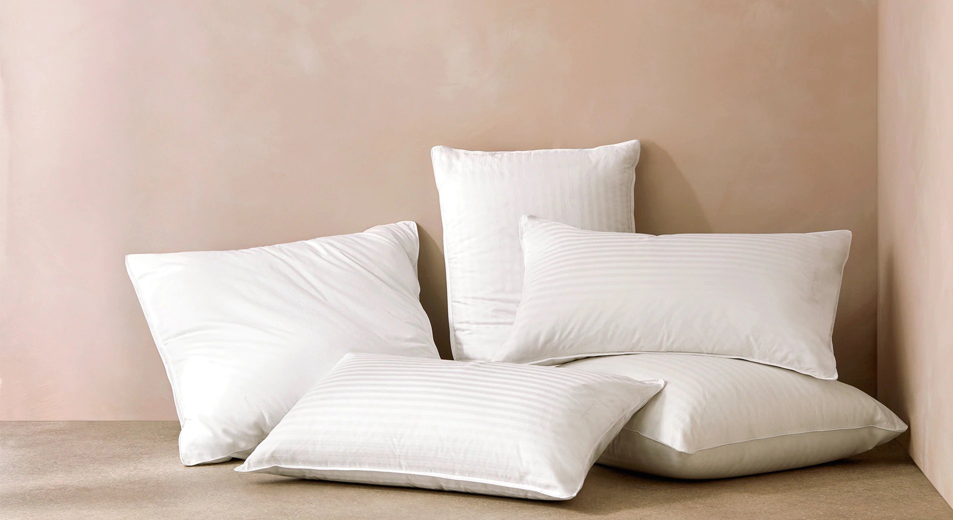 how to have a comfortable bed. five pillows of different sizes are stacked and placed against a beige wall.