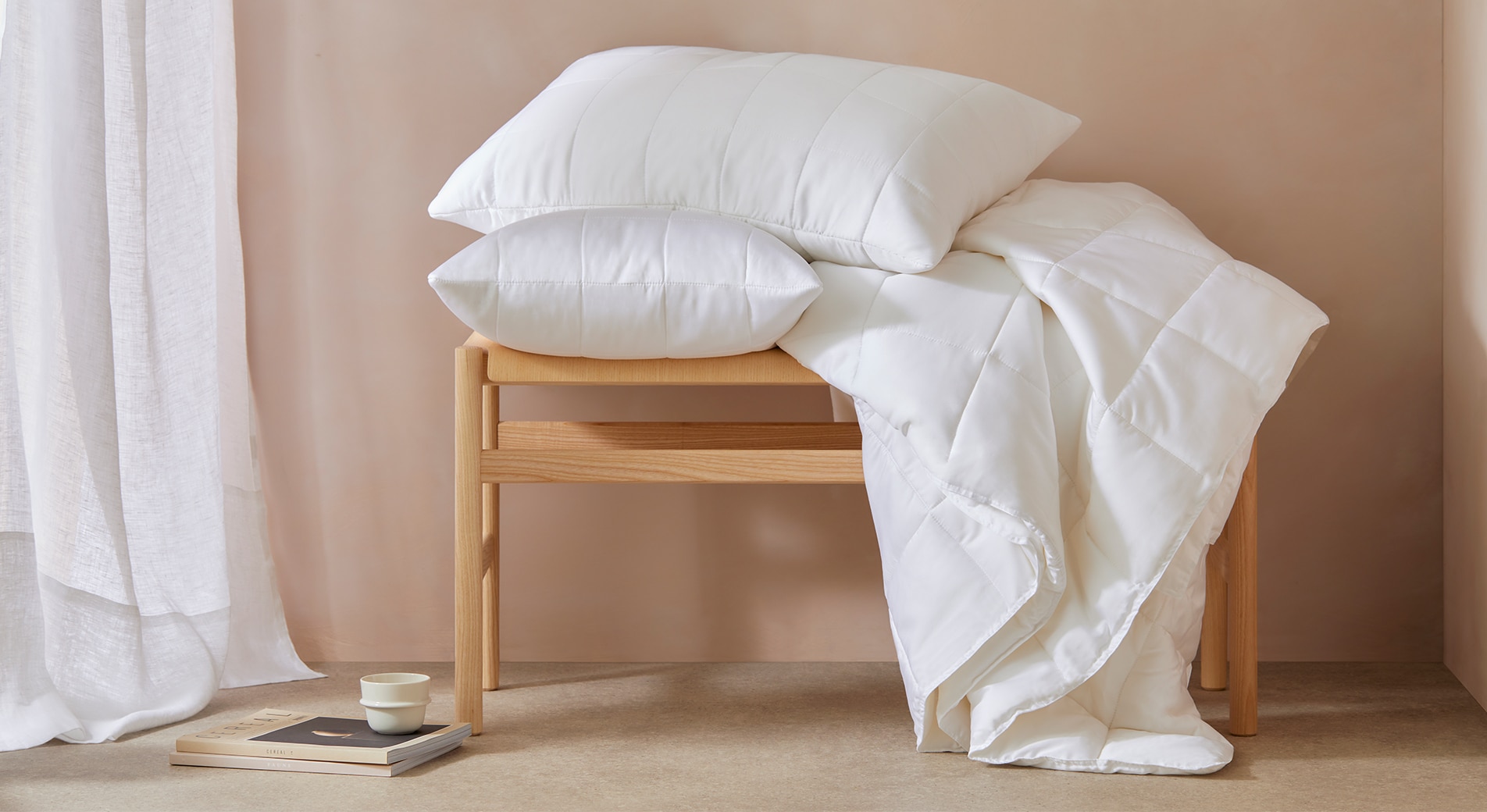 what makes a bed comfortable. two pillows and quilt are placed on a wooden bench in a biege room. there is a sheer curtain on the window, and magazine and mug on floor.