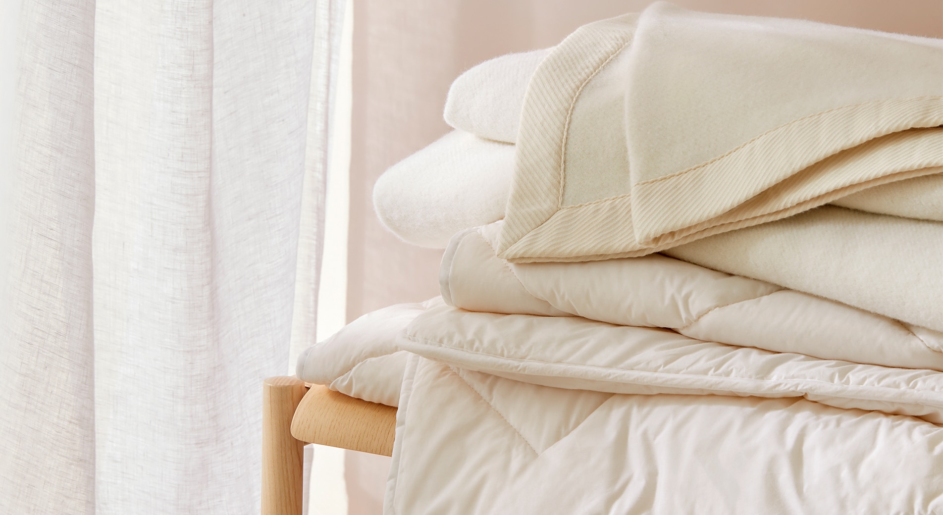 What should a bed include? Close up shot of wool blankets and quilt stacked on wooden bench. in the corner is sheer curtain.