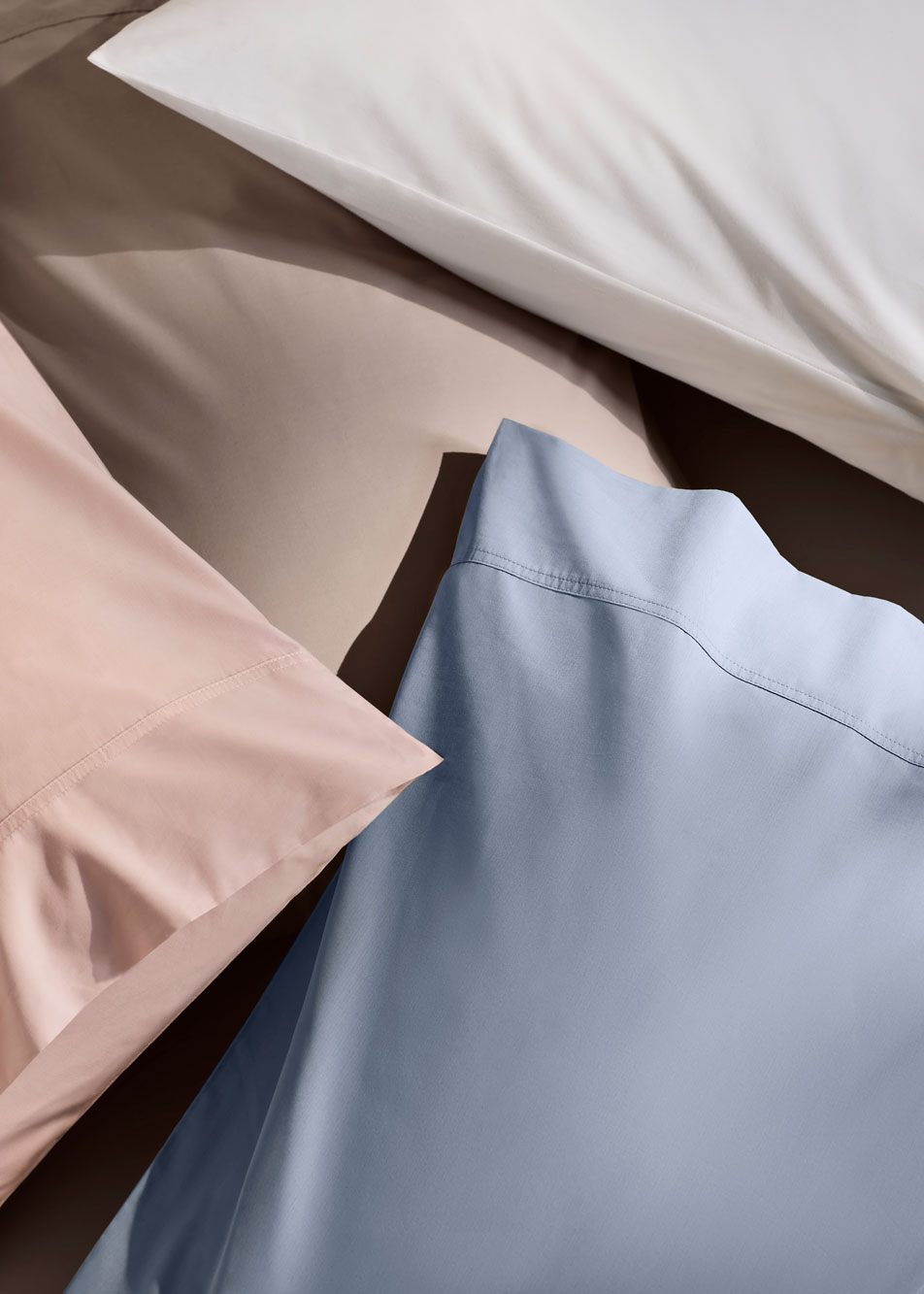 A close-up photo of a stack of pillows. Each pillow has a Byren pillowcase in a different shade: pink, white, blue and grey