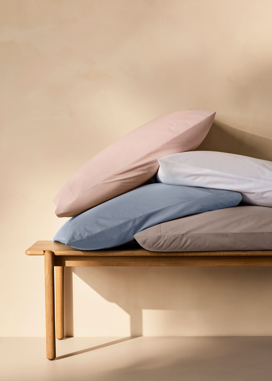 A stack of pillows sits on a timber bench. Each pillow has a Byren pillowcase in a different shade: pink, white, blue and grey
