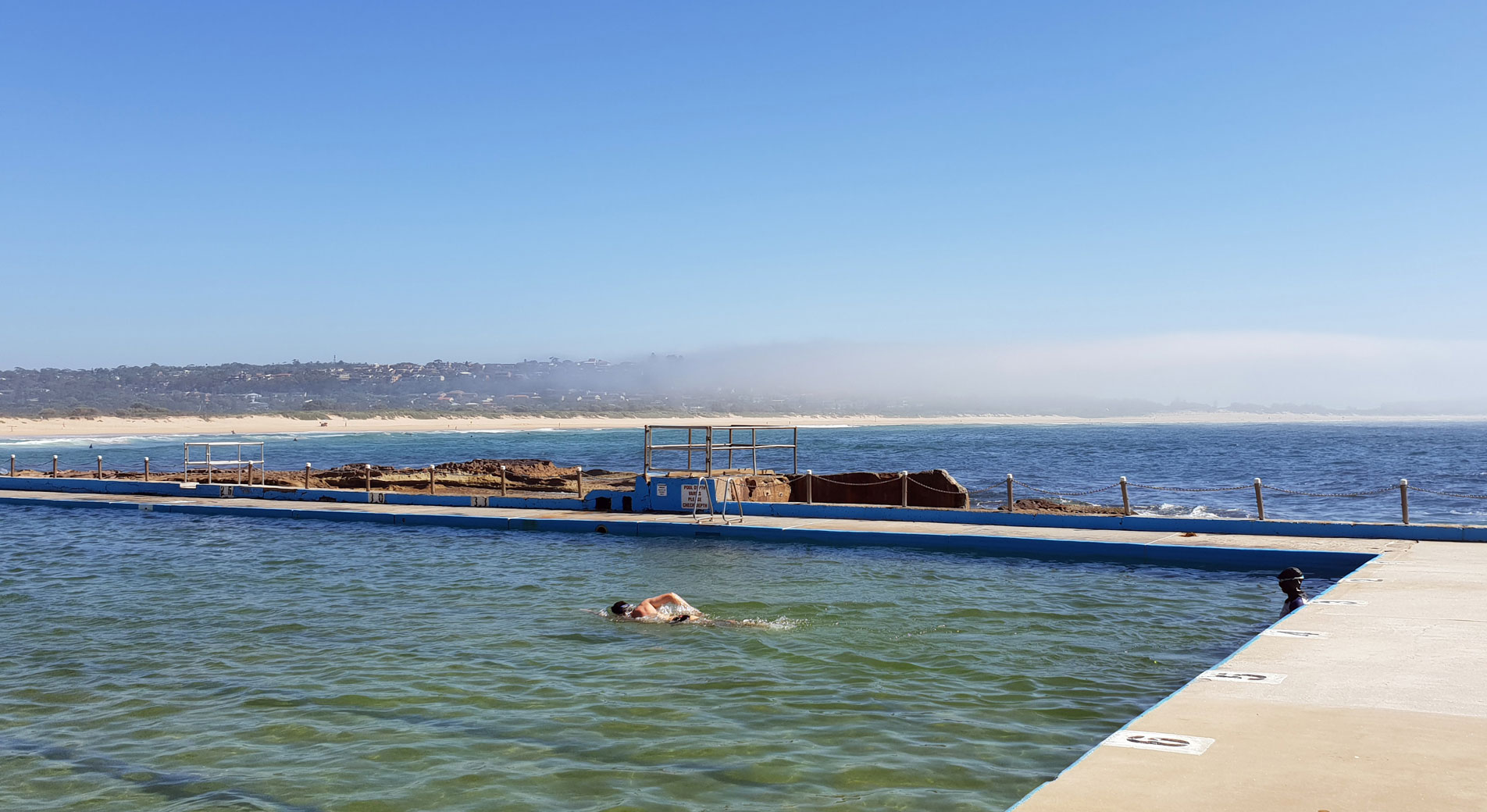 a shot of the outdoor swimming pool at dee why beach, with rocks and the stretch of sand in the background. in the foreground, a person swimps freestyle laps.