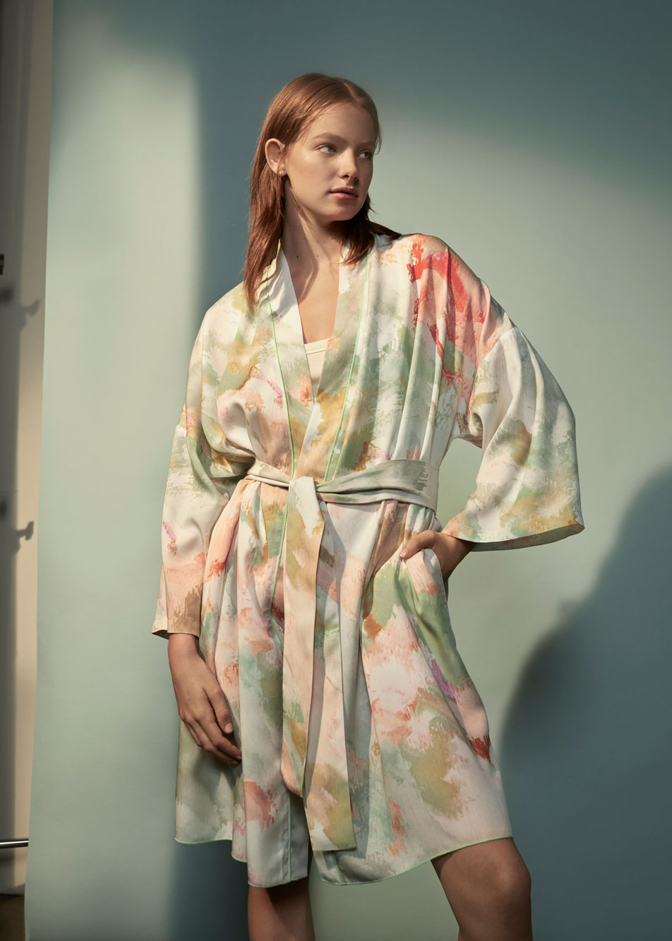 A young red headed woman standing in front of a pale green backdrop, wearing a Dovelake bath robe.