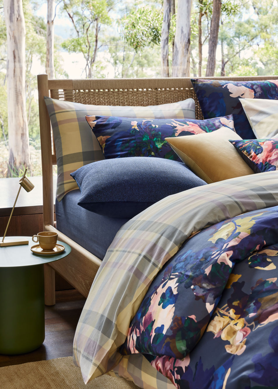 A bed sits in front of a window with a view of trees. The bed has layers of pillows and cushions, with two quilt covers in floral and check prints.