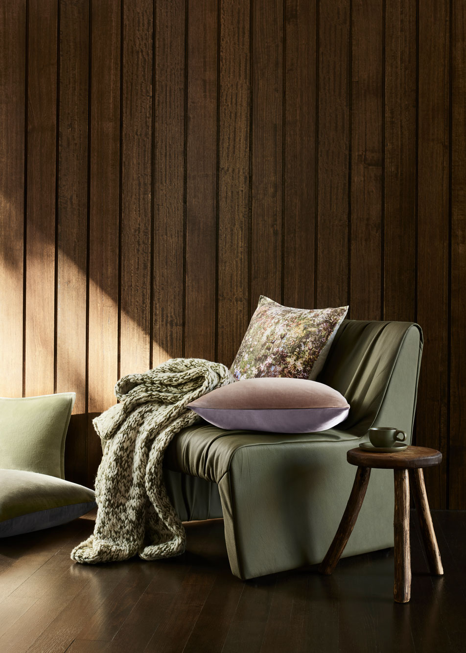 A green leather armchair in front of a timber wall. On top of the chair is two cushions and a knit throw.