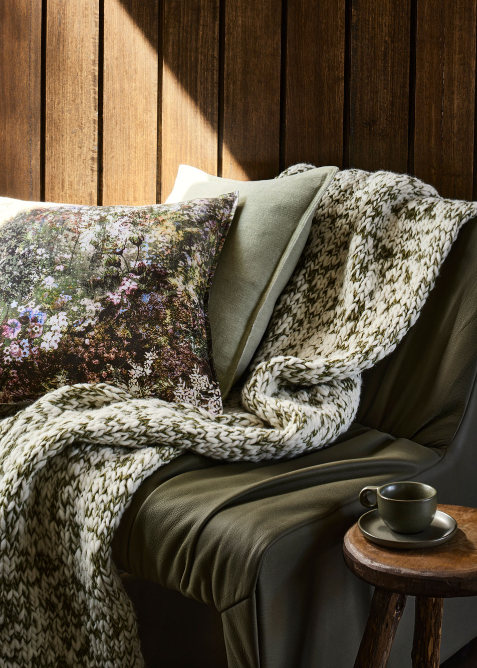 A green and white knit throw sits on top of a deep green leather chair, next to a timber side table. Multicoloured cushions are scattered around.