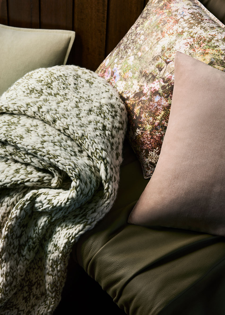 A green and white knit throw sits on top of a deep green leather chair. Multicoloured cushions are scattered around.