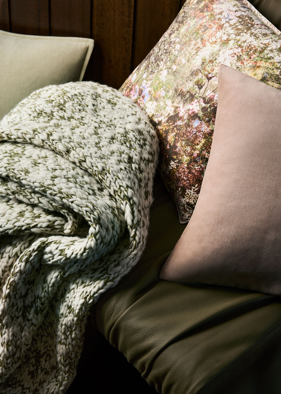 A green and white knit throw sits on top of a deep green leather chair. Multicoloured cushions are scattered around.