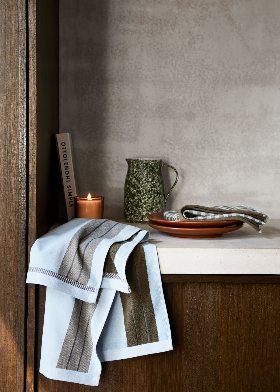 A blue striped tea towel draped over a counter top. Beside it sits a lit candle, cook book and ceramic water jug.