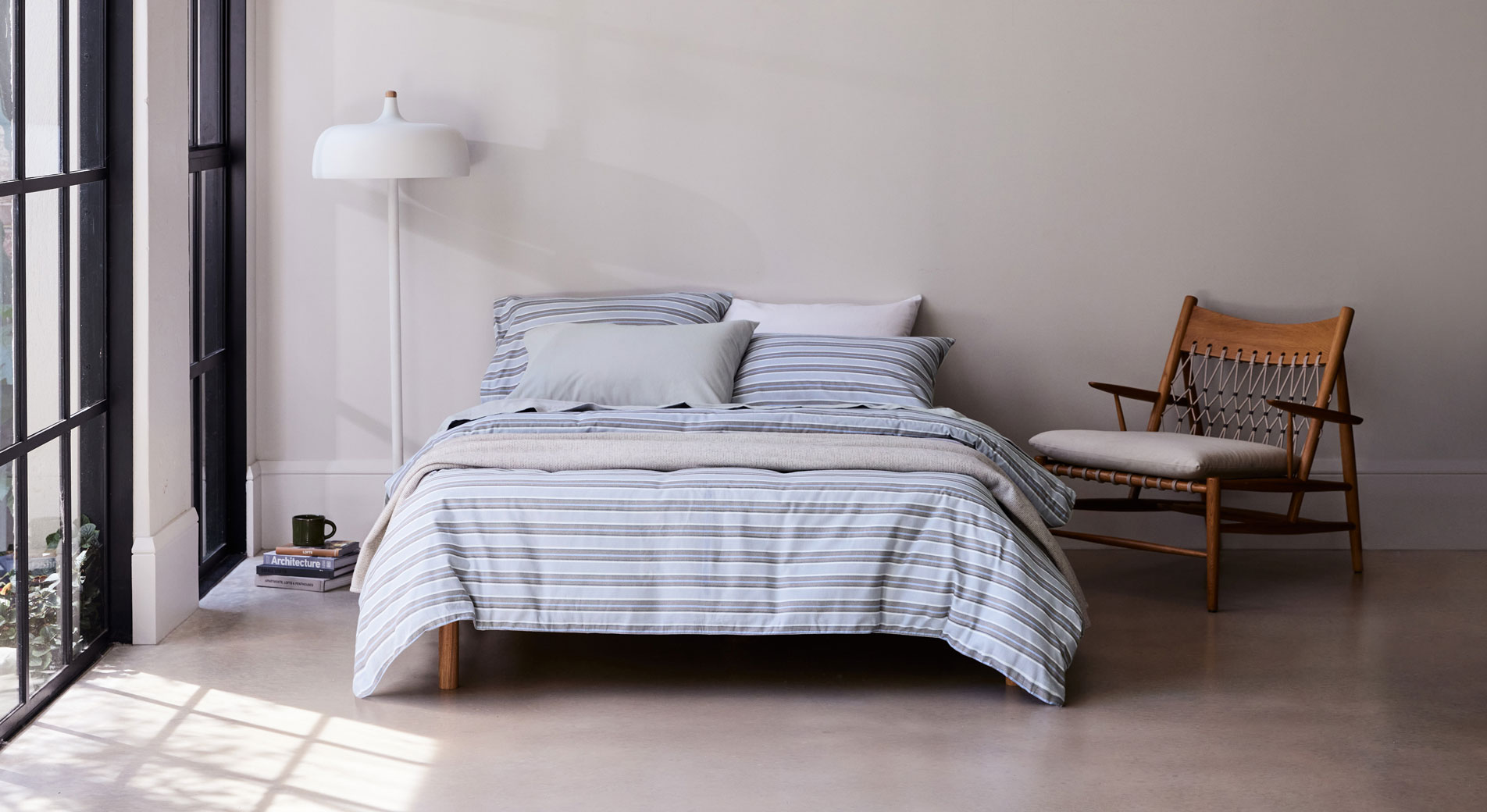 A bed dressed in blue striped flannelette bedding sits in a modern bedroom with concrete floors and French windows. Next to the bed is a wooden armchair, a white lamp and a small stack of books with a mug sitting on top.