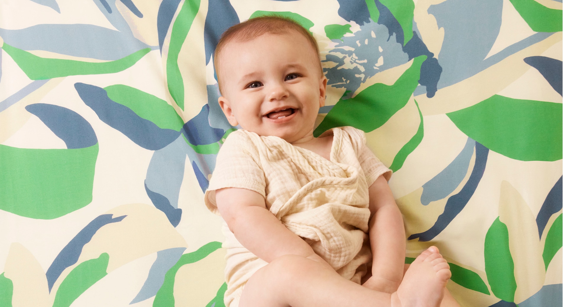 baby with gummy smile smiles up at camera. wearing neutral clothing, against sheridan mysie blanket with green blue and biege floral pattern in background