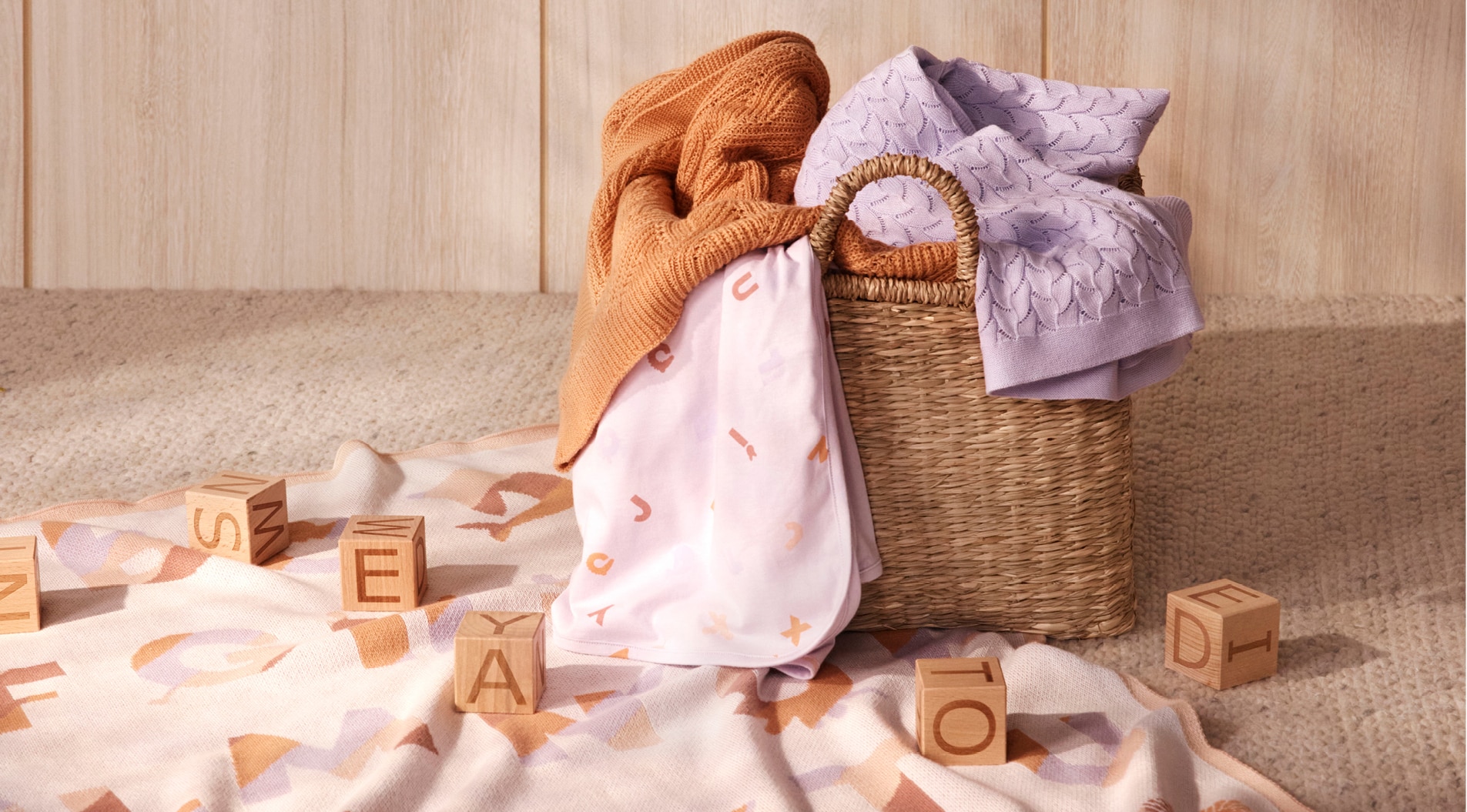 image of baby nursery. letter blocks are scattered across alphabet blanket laid on floor. woven basket contains sheridan blankets, including orange and lilac ones.