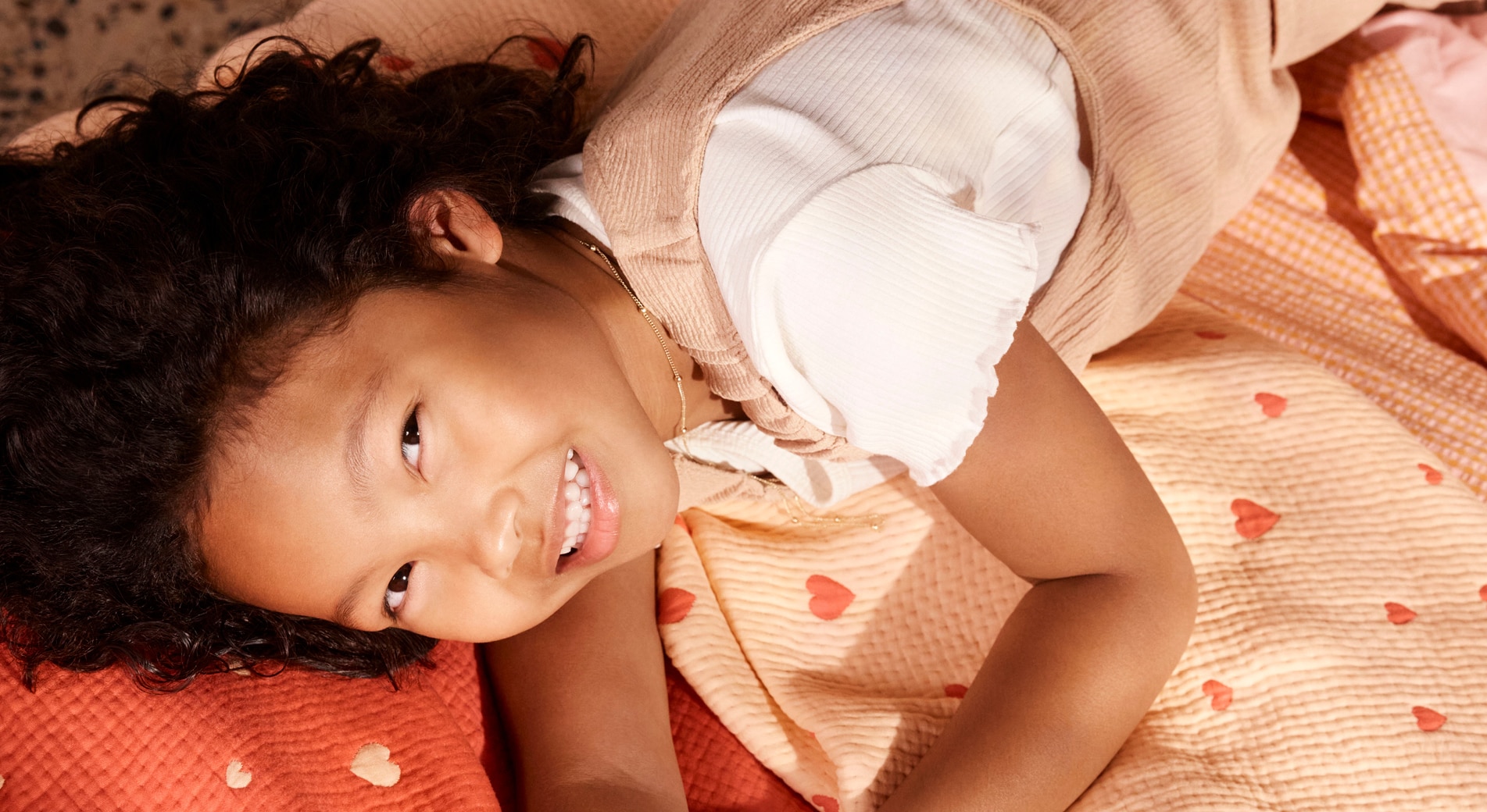 landscape photo of young girl lying on sheridan bedding. she wears a white top and pink coveralls, lying on quilted peach and red bed covers, quilted with hearts