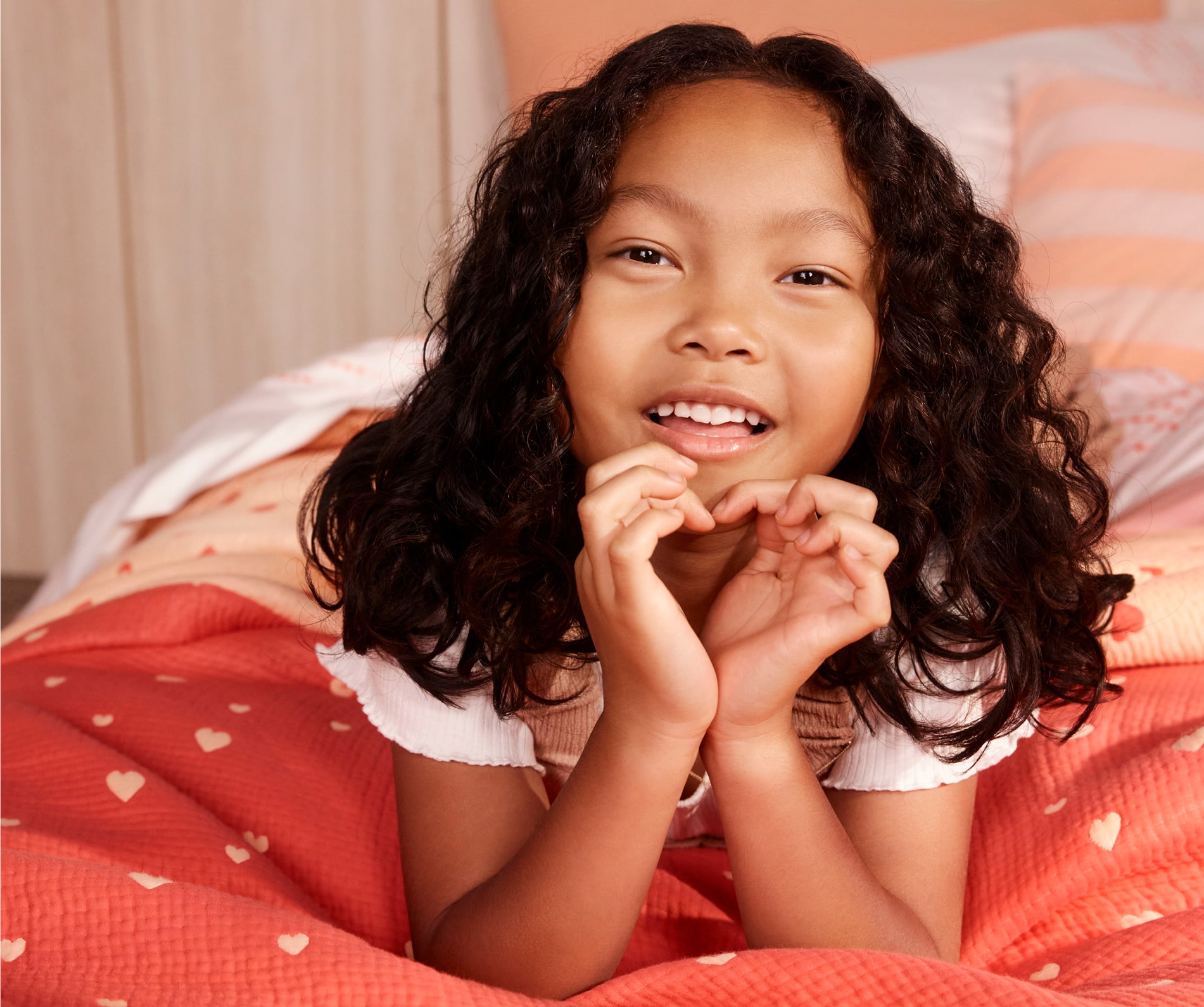 young girl lies on a bed, on her stomach with her elbows propped up, smiling at the camera and making the shape of a heart with her hands. bed has been styled with quilted bed cover, red and peach with little hearts.