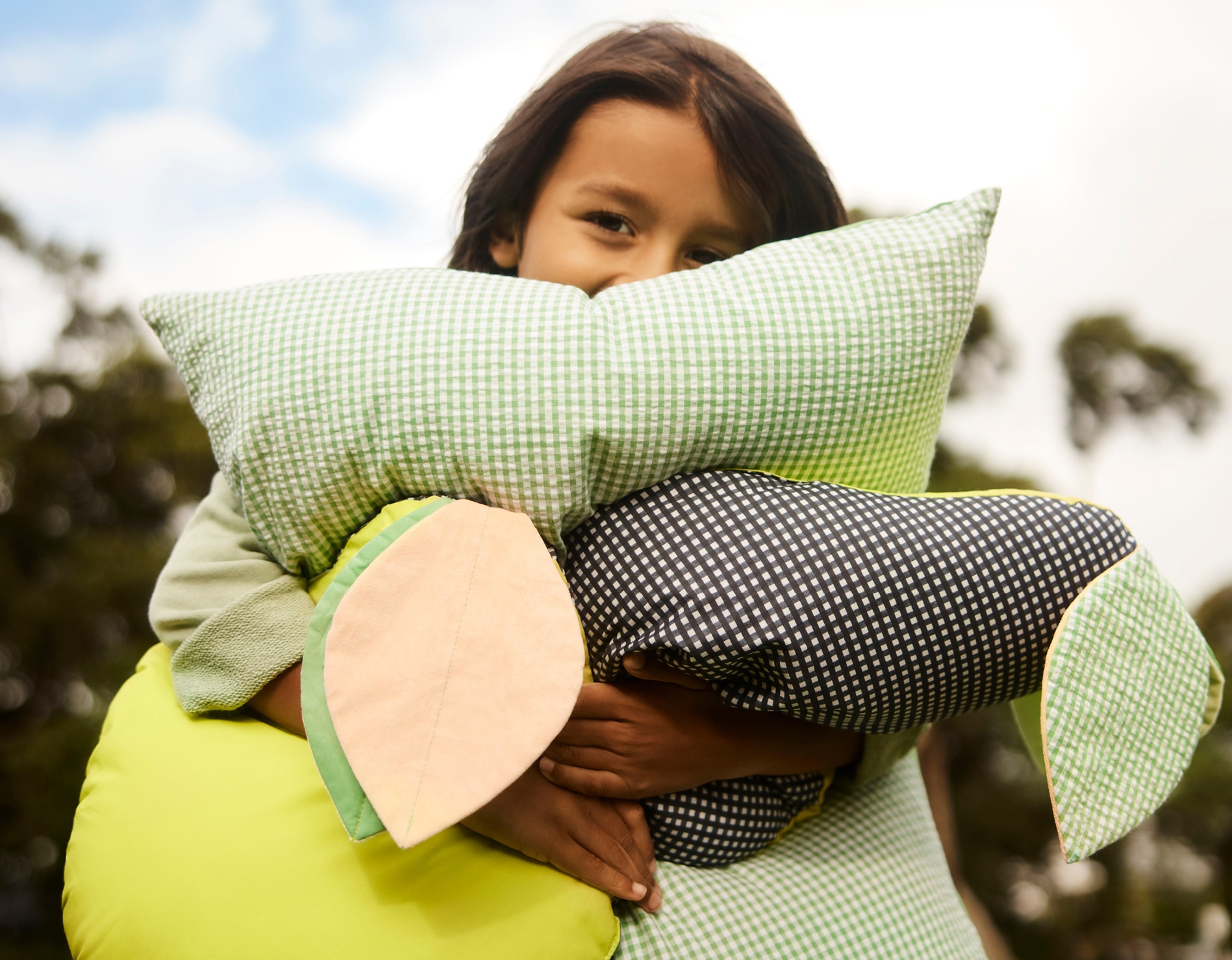 young boy, clutching a pile of decorative sheridan kids cushions, stands outside. half his face is shyly hiding behind the cushions, though you can tell he's smiling. background of trees, blue sky and clouds, out of focus.