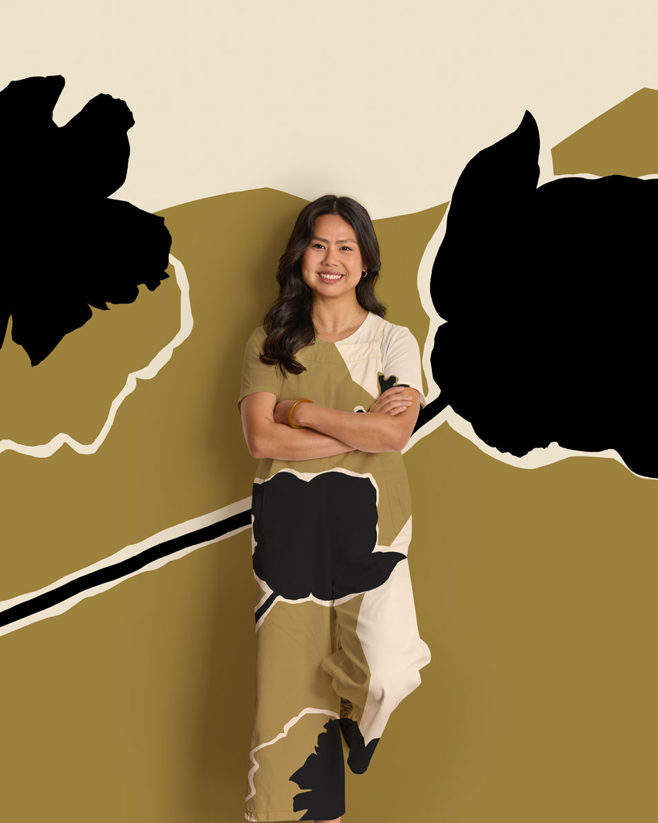 A woman stands, smiling in front of an abstract floral print background. Her jumpsuit is in the same matching pattern.