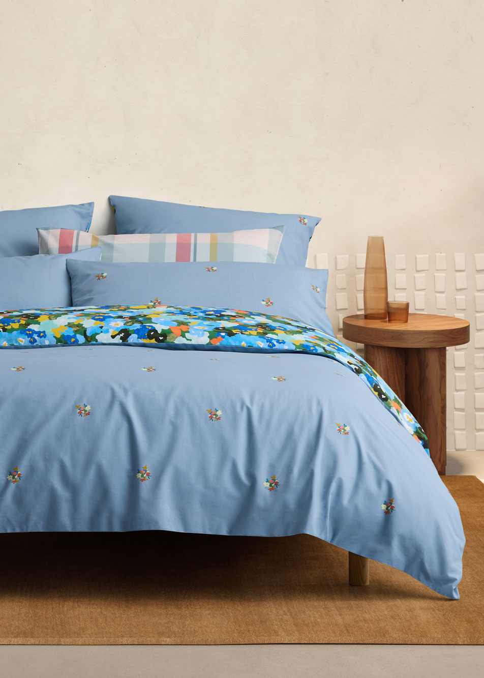 A blue floral bed sits in a beige bedroom. Next to the bed is a timber side table with a glass carafe.