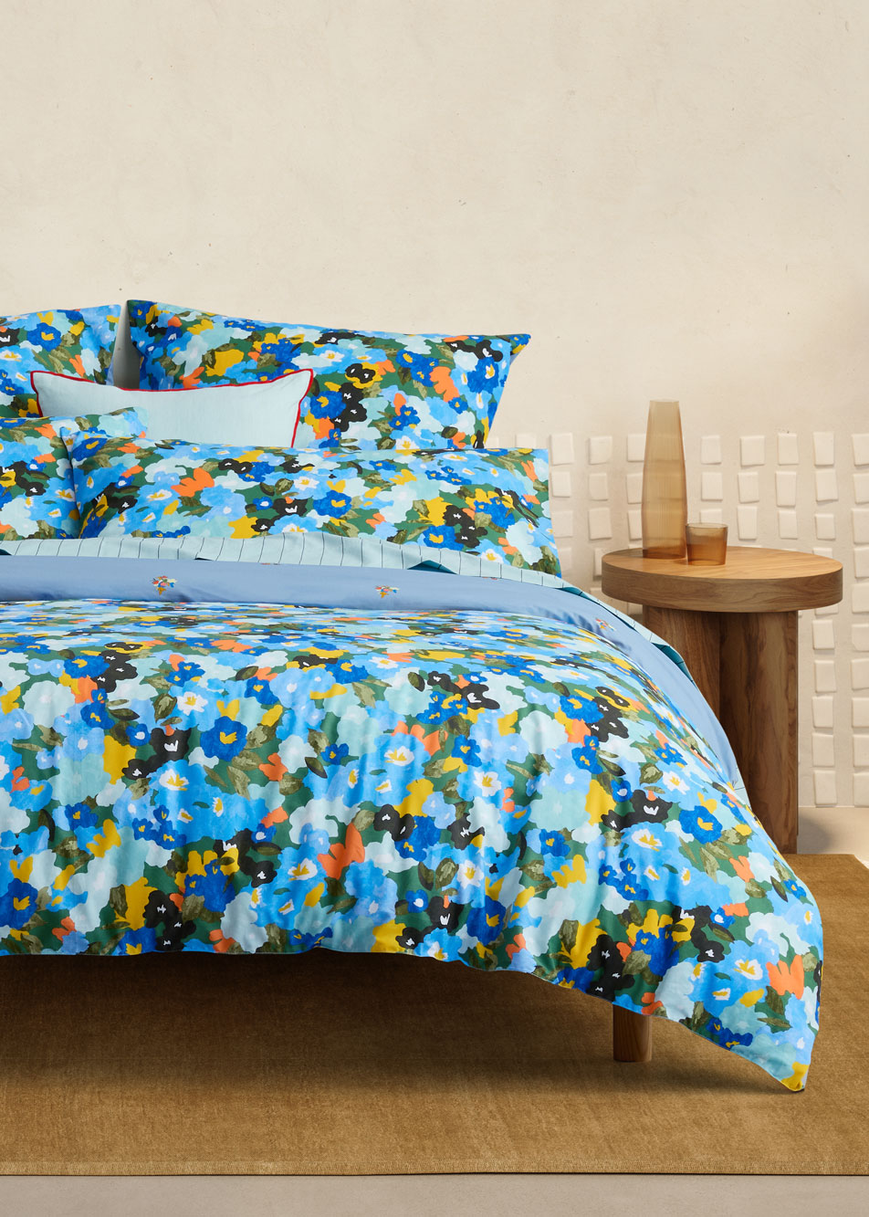 A colourful floral bed sits in a beige bedroom. Next to the bed is a timber side table with a glass carafe.
