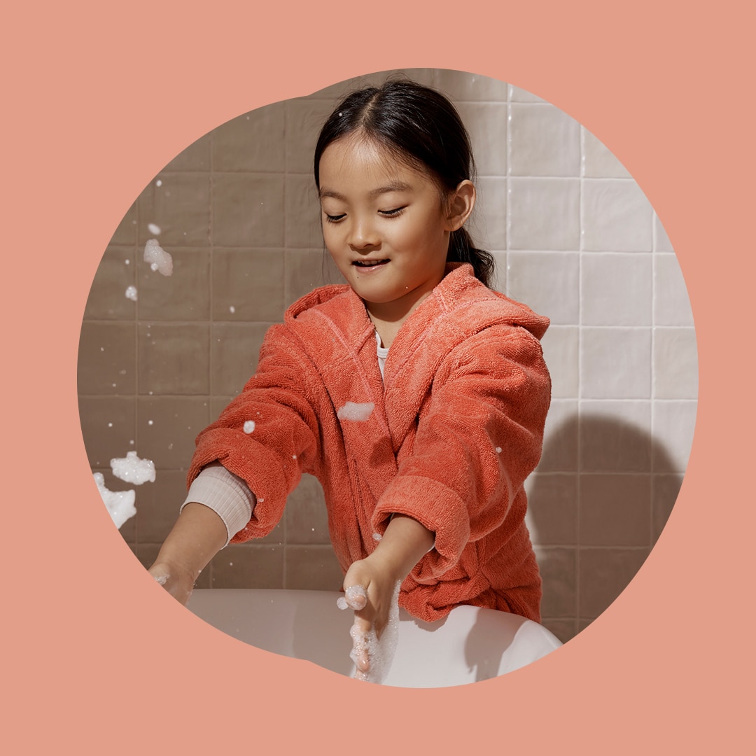little girl with black hair wears pyjamas and bathrobe while she leans over bathtub, throwing bubbles in the air while smiling.