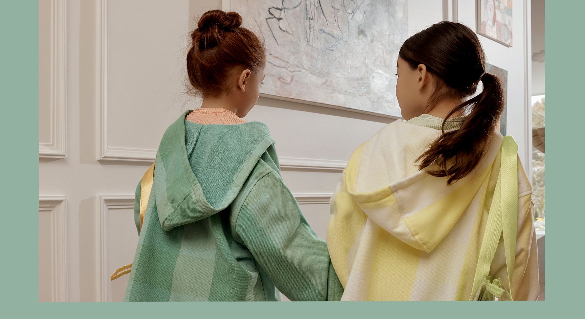 two little girls wearing tessy ponchos walk away from the camera holding hands. they have bags over their shoulder, like they're going to the beach.