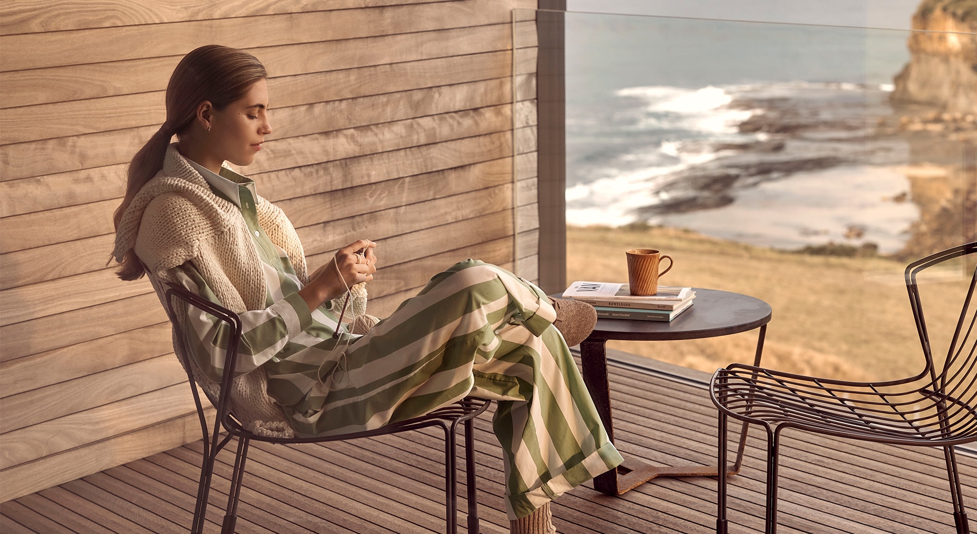 woman in striped green and white loungewear set sits on a balcony outside, with wooden wall and wooden floor, and glass railing. she has a view of grassy knoll, leading down to rocky cliff front and ocean.