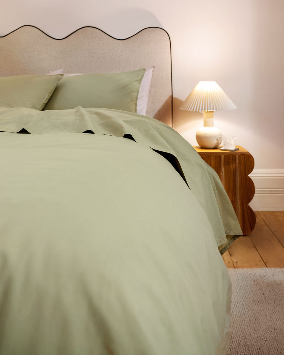 A close-up image of a green bed in a neutral bedroom. Beside the bed is a sculptural timber side table with a white lamp.