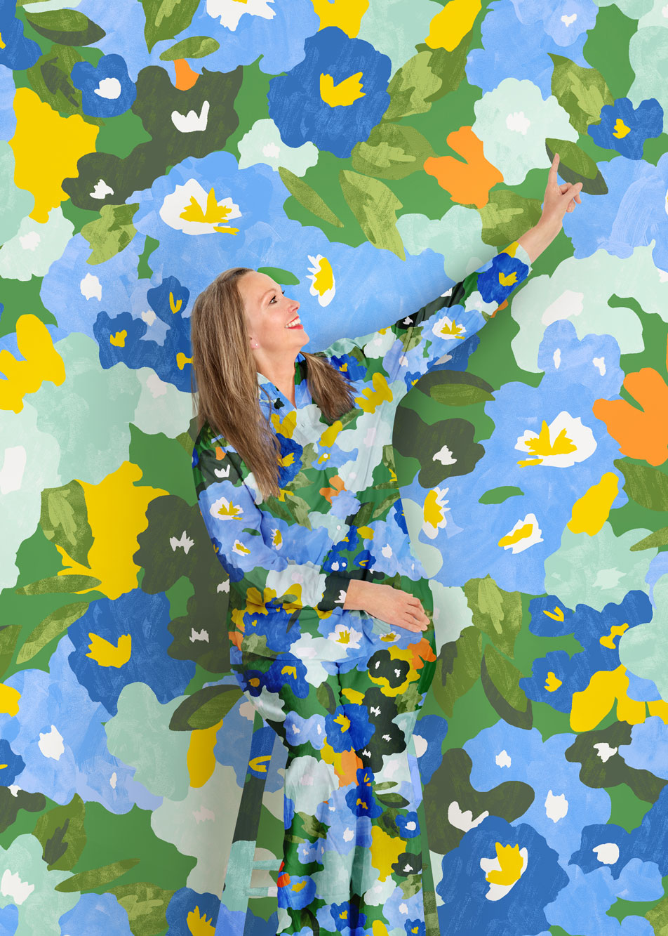 A woman sits, smiling and pointing up at a vivid floral print background. Her shirt and pants are in the same matching pattern.