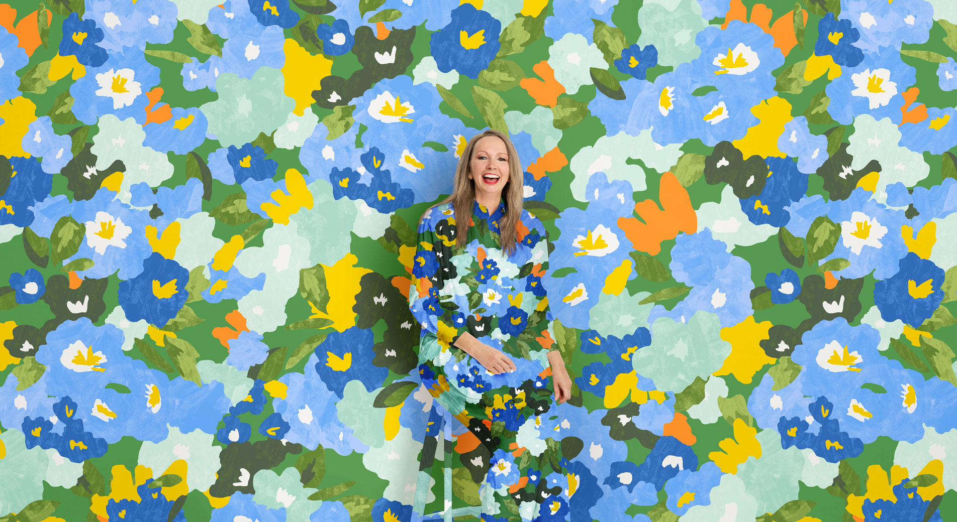A woman sits, smiling in front of a vivid floral print background. Her shirt and pants are in the same matching pattern.