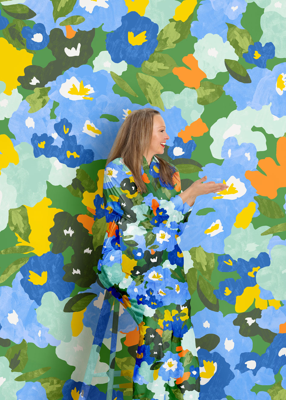 A woman sits, smiling in front of a vivid floral print background. Her shirt and pants are in the same matching pattern.