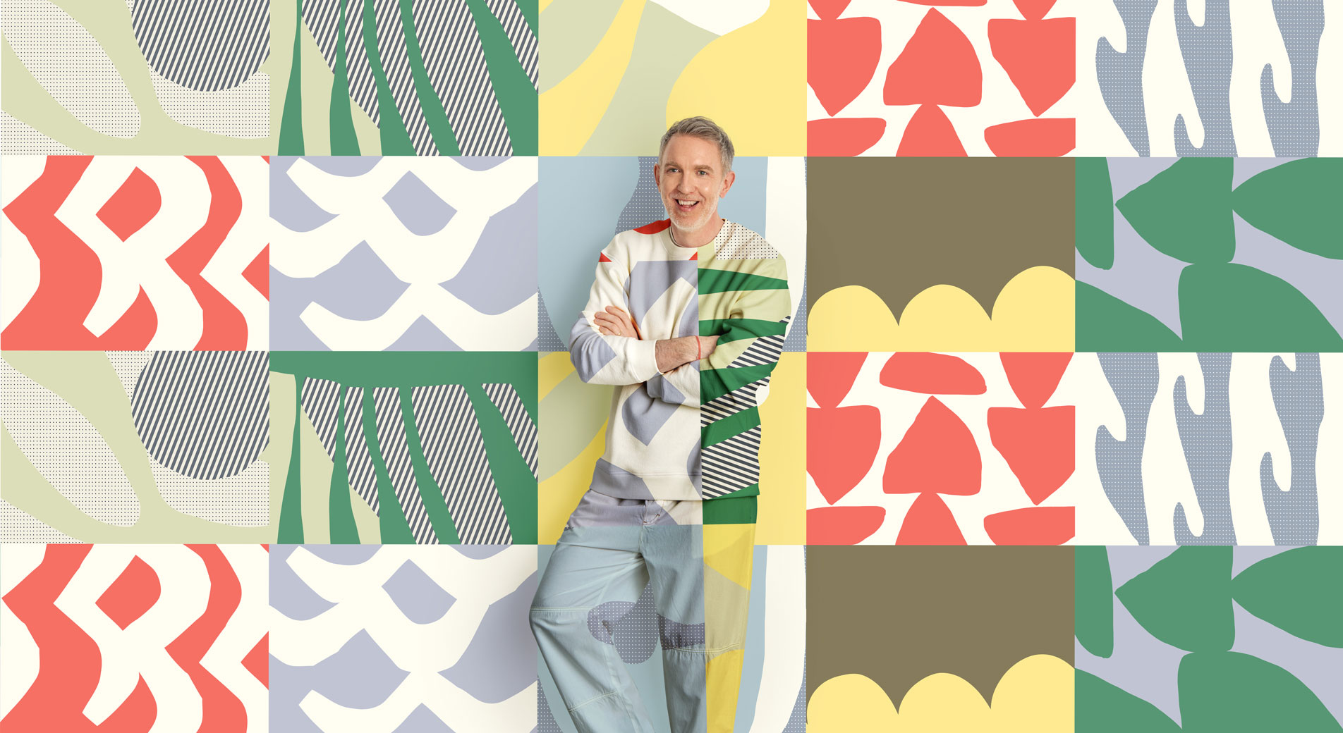A man stands, smiling in front of a colourful geometric print background. His shirt and pants are in the same matching pattern.