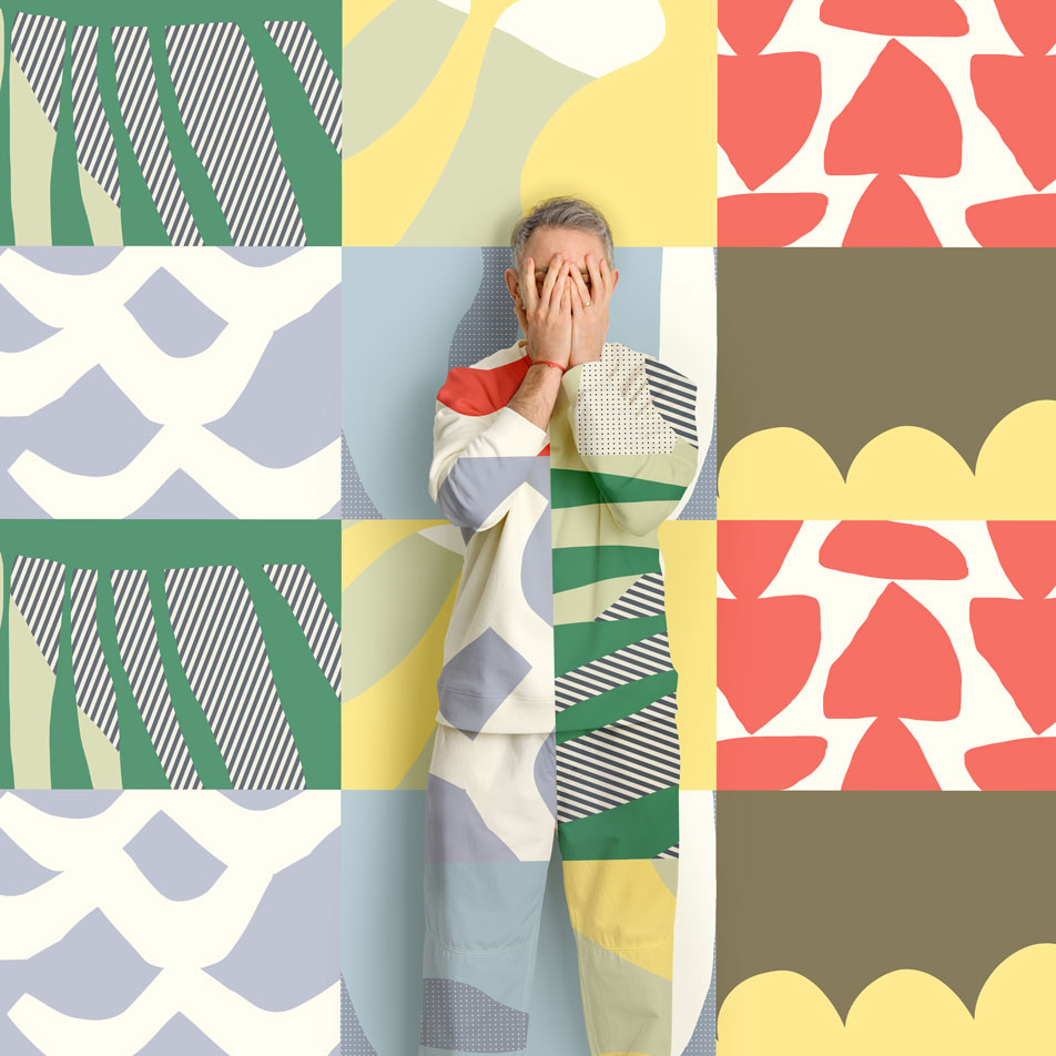 A man standing in front of a geometric print background, playfully covering his face with his hands. His clothes are the same geometric print as the backdrop.