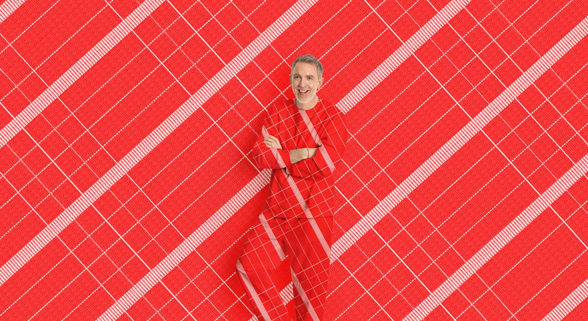 A man stands, smiling with his arms crossed in front of a red and white check print background. His shirt and pants are in the same matching pattern.