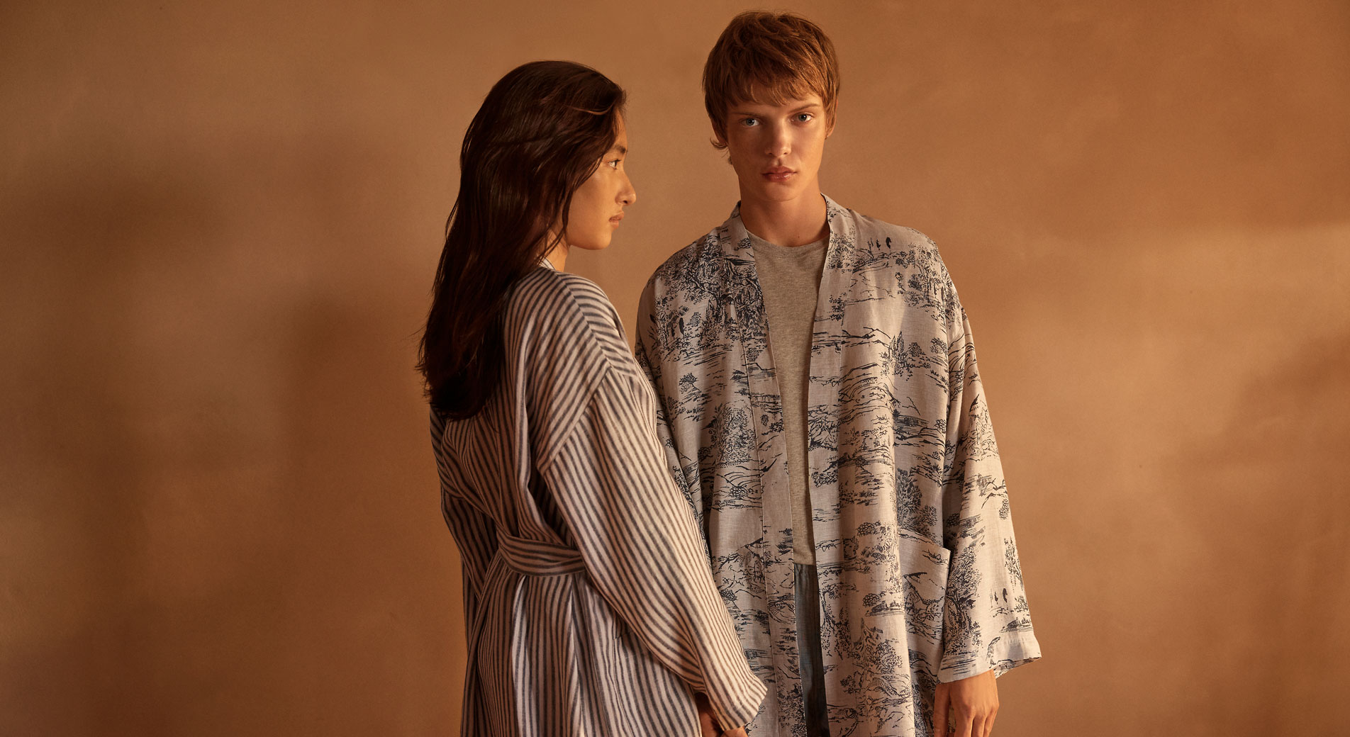 Young man and woman stand in front of a brown painted wall, both wearing stylish linen robes 