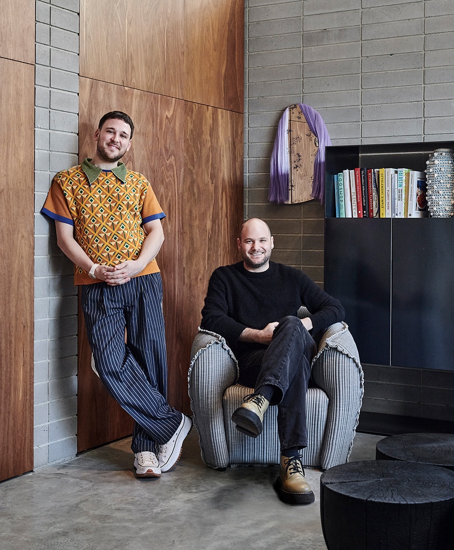 james vivian and husband ben asakoff pose in corner of their living room. james leans against wall, wearing navy striped pants, patterned autumnal polo, sneakers. ben sits on textured chair, one leg resting atop the other, wearing all black