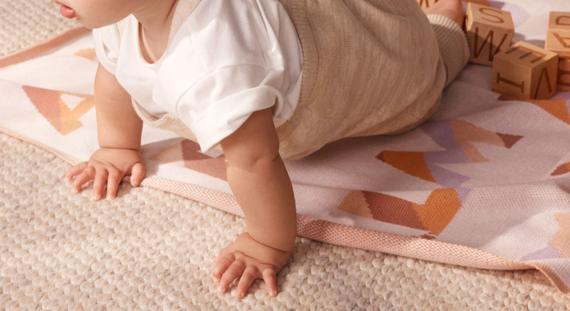 cropped landscape shot of baby enjoying tummy time on jute rug, with sheridan aleph blanket beneath him. he has his mouth open and looks alert, wearing a white tee with rolled up sleeves and beige joggers.