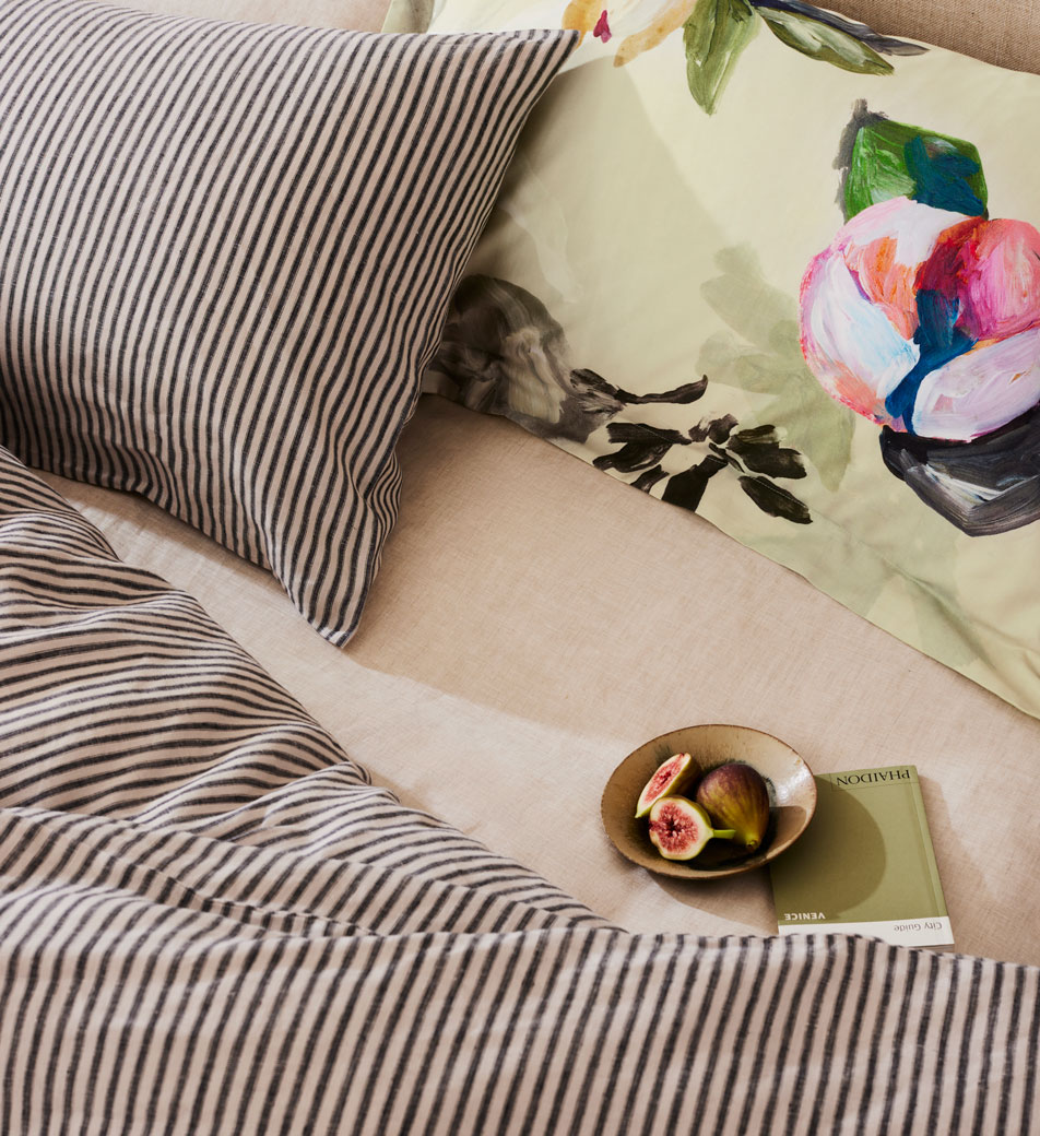 Mixing stripes and florals. Close up of a neutral bed with grey striped sheets and a green floral pillow. On top sits a bowl of figs and a small green notebook 