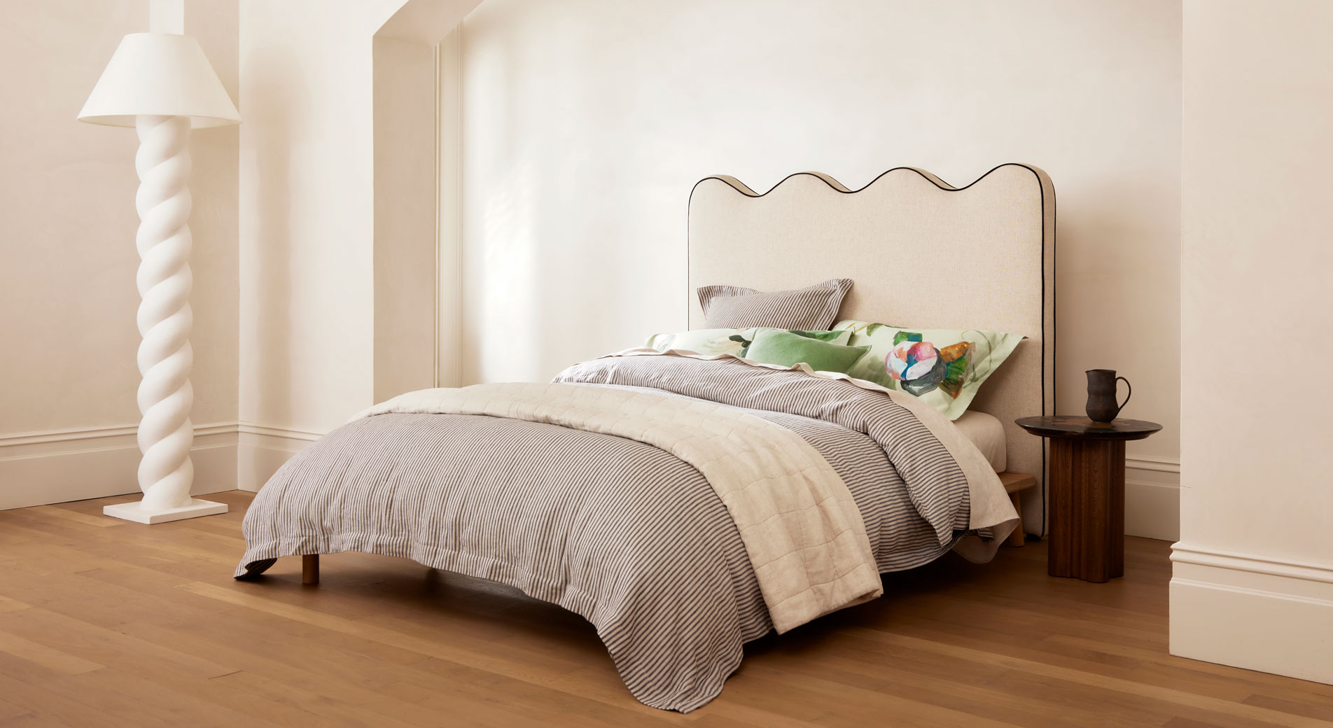 Industrial style bedroom. A grey striped bed sits in a room with white walls, timber flooring and a white sculptural spiral lamp.