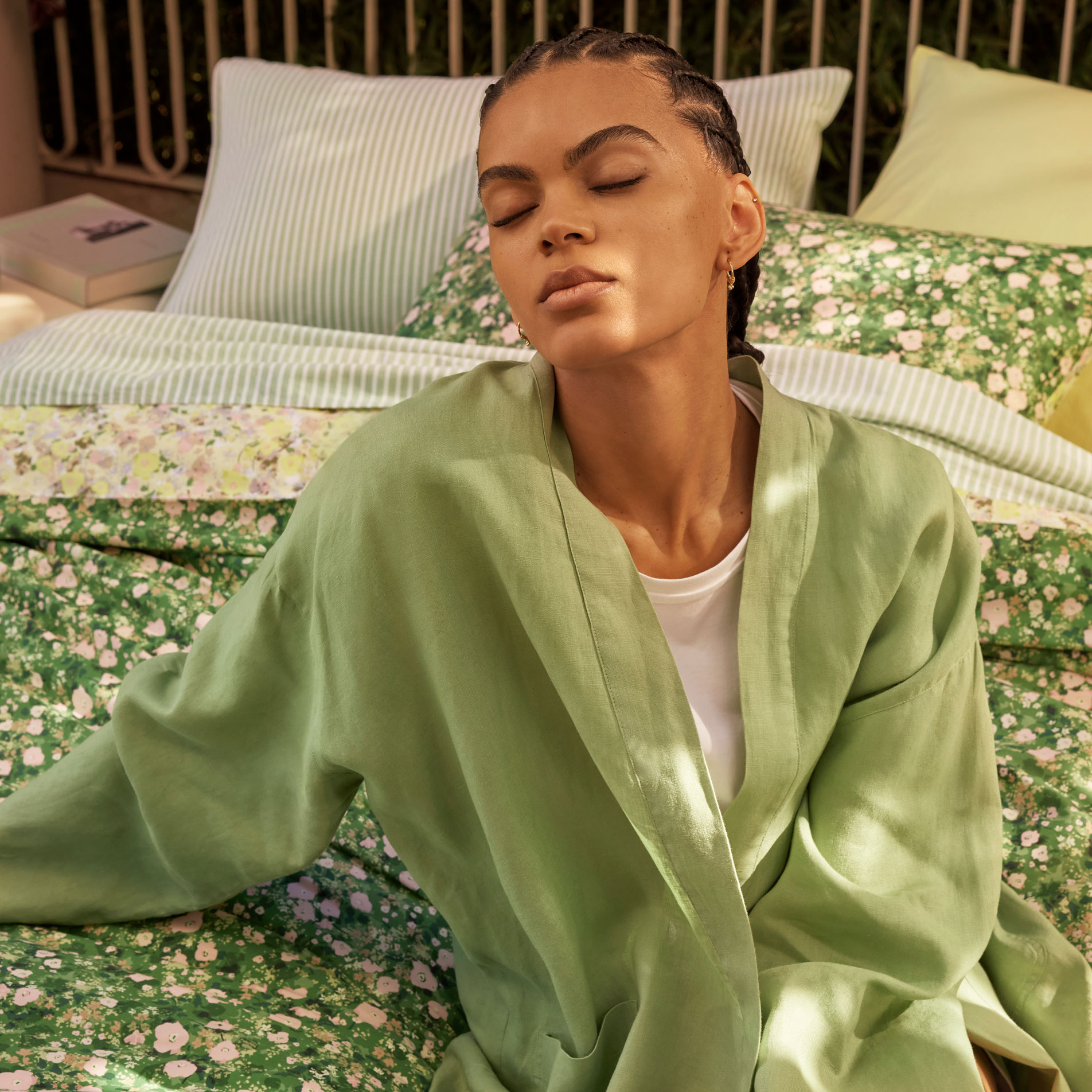 A young woman wearing a green linen robe and braided hair lounges on a floral print bed outside on a balcony, with dappled sun shining on her face