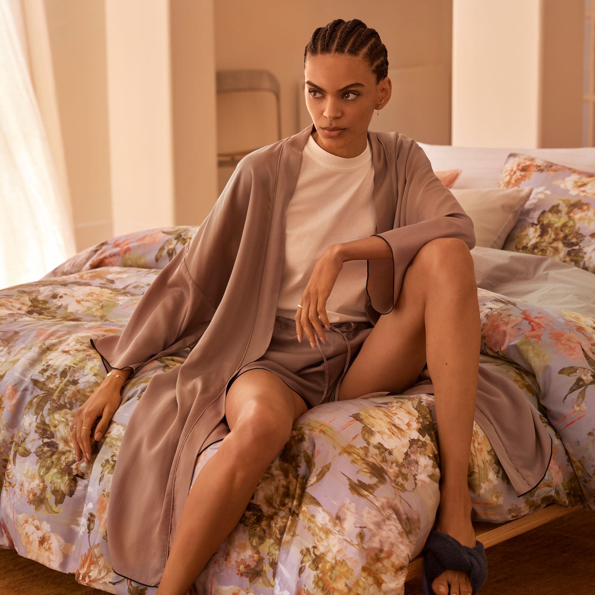 A young woman wearing silky loungewear and braids sits on the corner of a floral print bed, by a window in a neutral-toned room