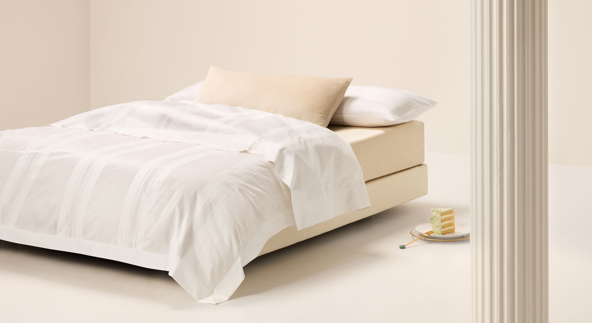 A bed sitting in an empty white room. The bed is dressed in Millennia bedding. On the floor next to the bed is a slice of cake on a plate.