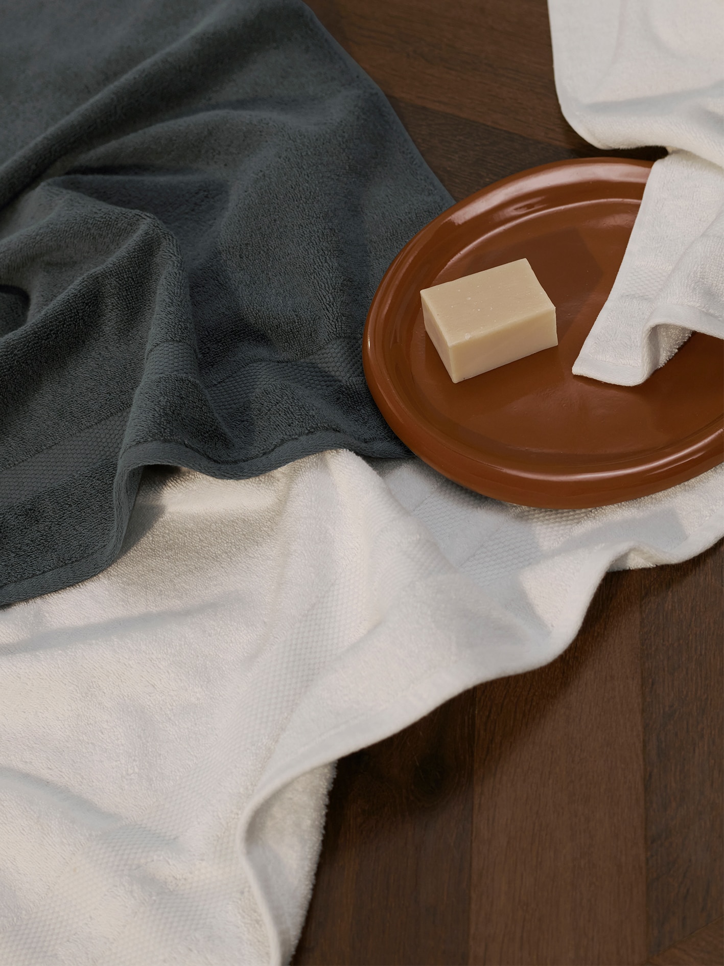 styled towel flat lay. meridian towels in blue and white laid on polished wooden floor. on a brown ceramic plat is a square of soap.