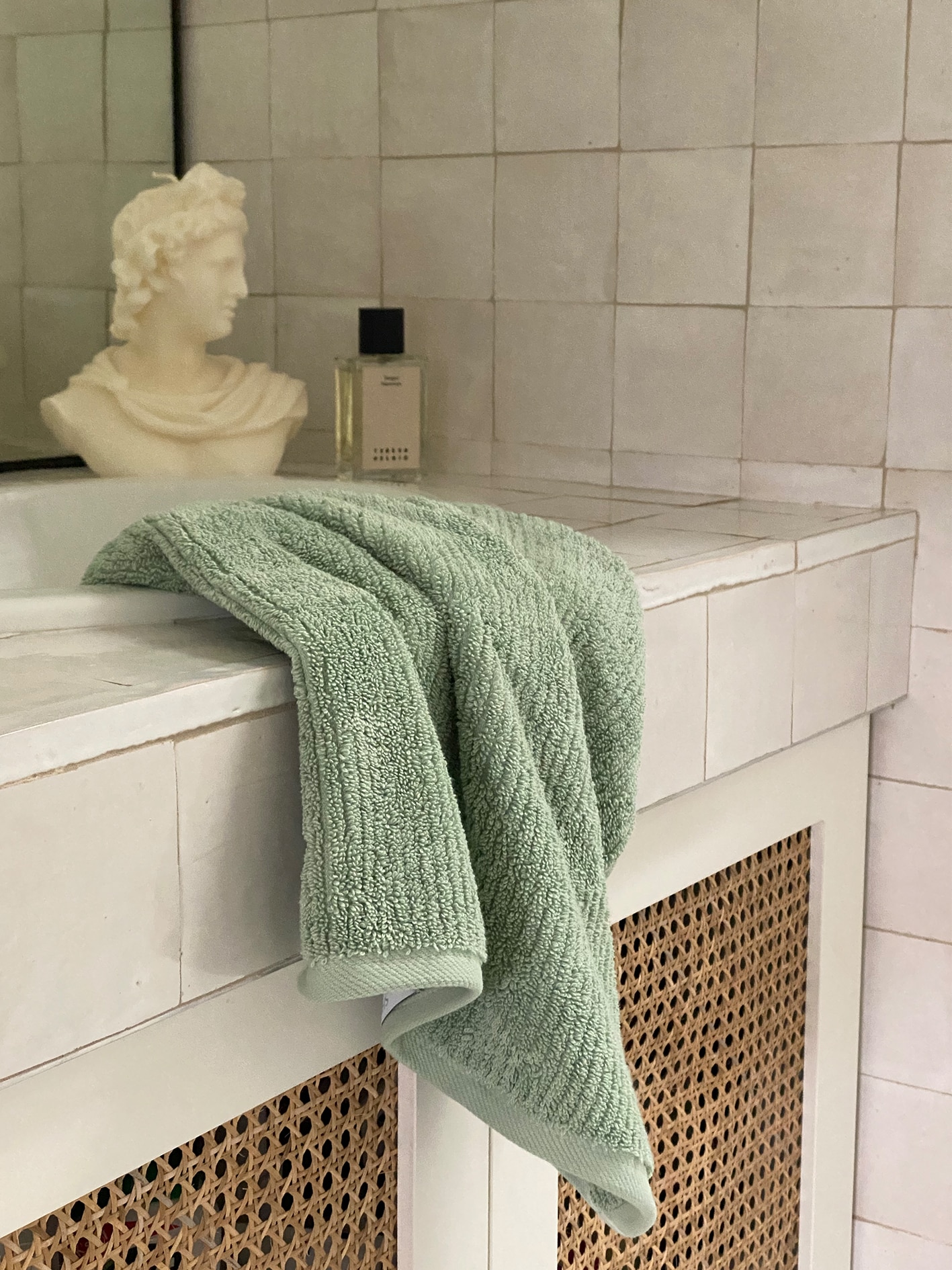 close up of bathroom counter. wicker doors underneath, marble tiles on top. mint towel lays atop counter into sink. in the background is a candle bust, and perfume.