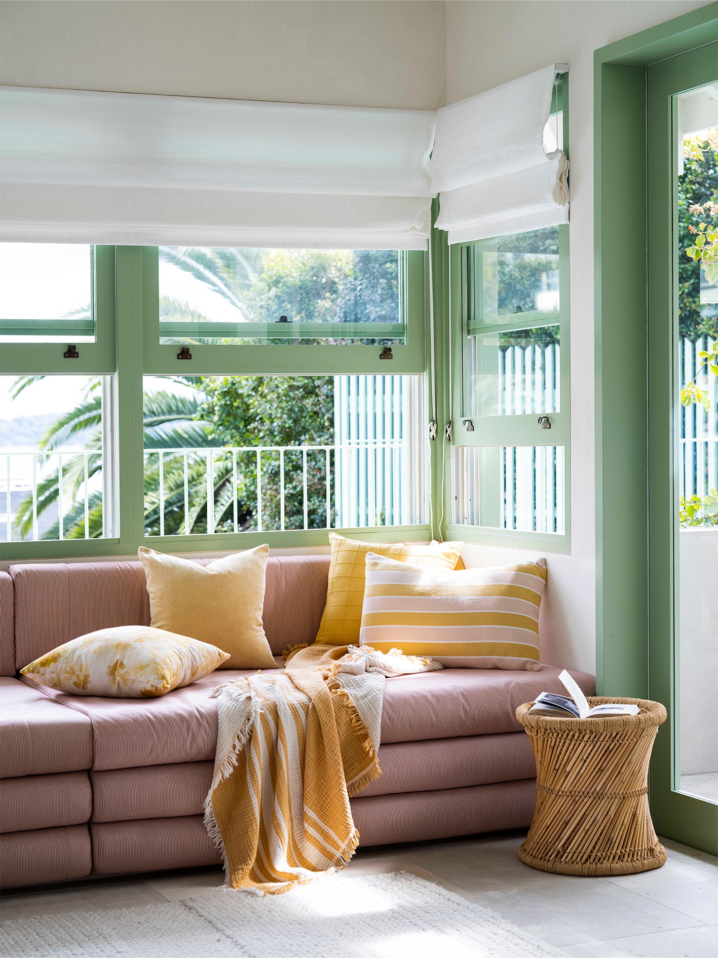 pink couch layered with gold and yellow cushions and throws. windows in abckground with green awnings. biege floor, on it sits a wooden stool/feature side table.