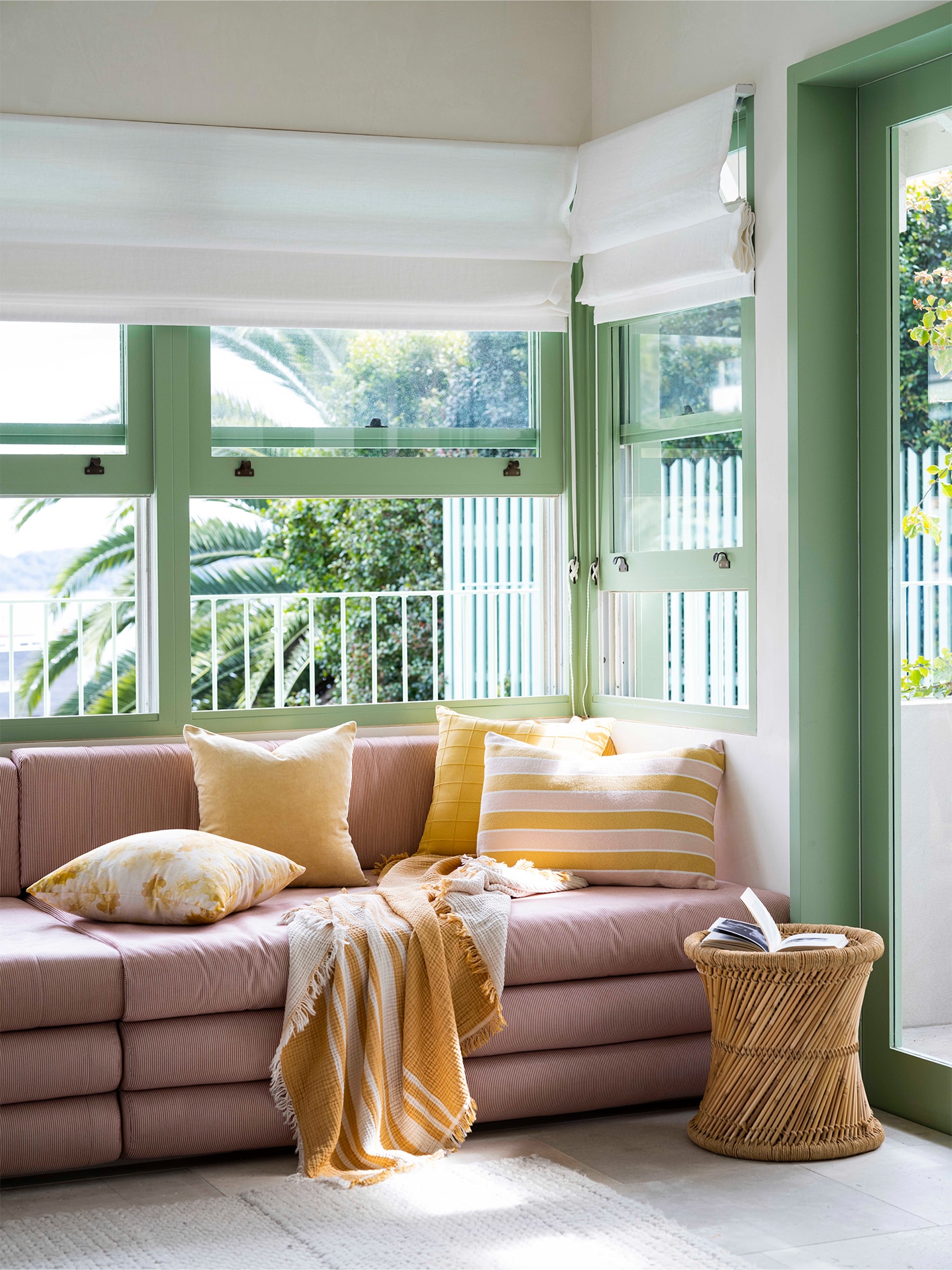 pink couch layered with gold and yellow cushions and throws. windows in abckground with green awnings. biege floor, on it sits a wooden stool/feature side table.