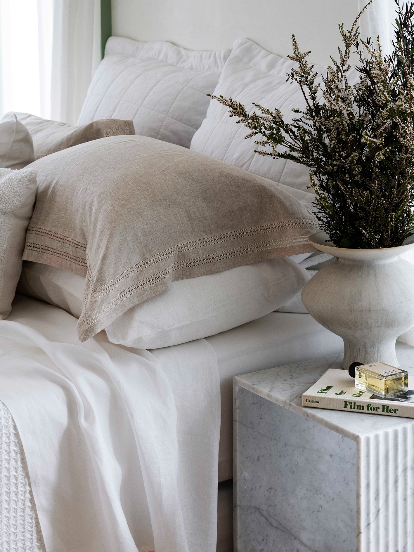styled bed. six cushions stacked neatly, in shades of white and flax. marble bedside table sits next to bed, with a book and plant in marble pot.