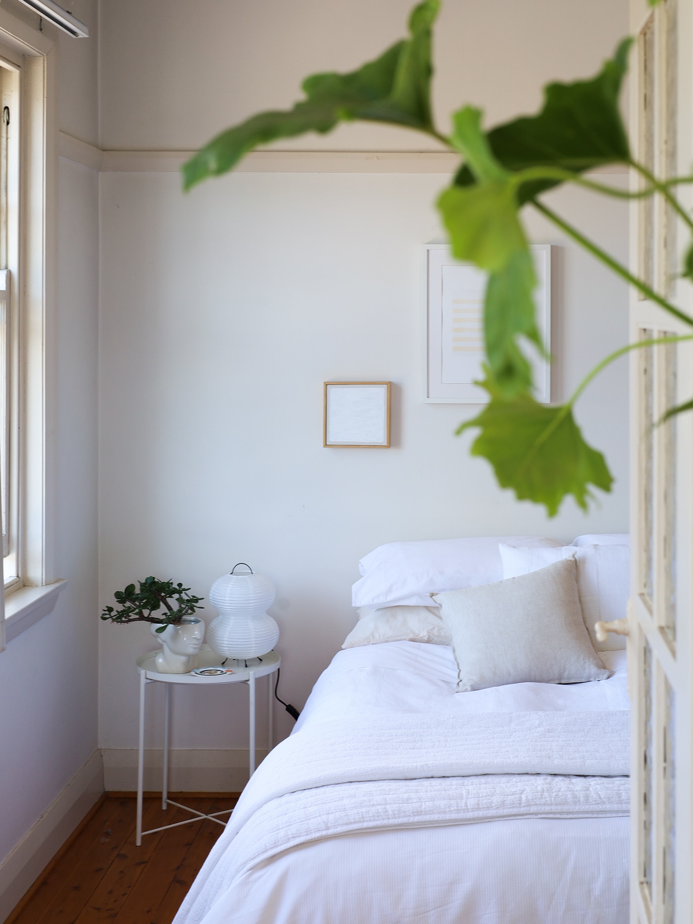 vertical shot through door into bedroom. window on left hand side. white bed styling with flax coloured cushion and sheet. green plant pokes through at front of image.