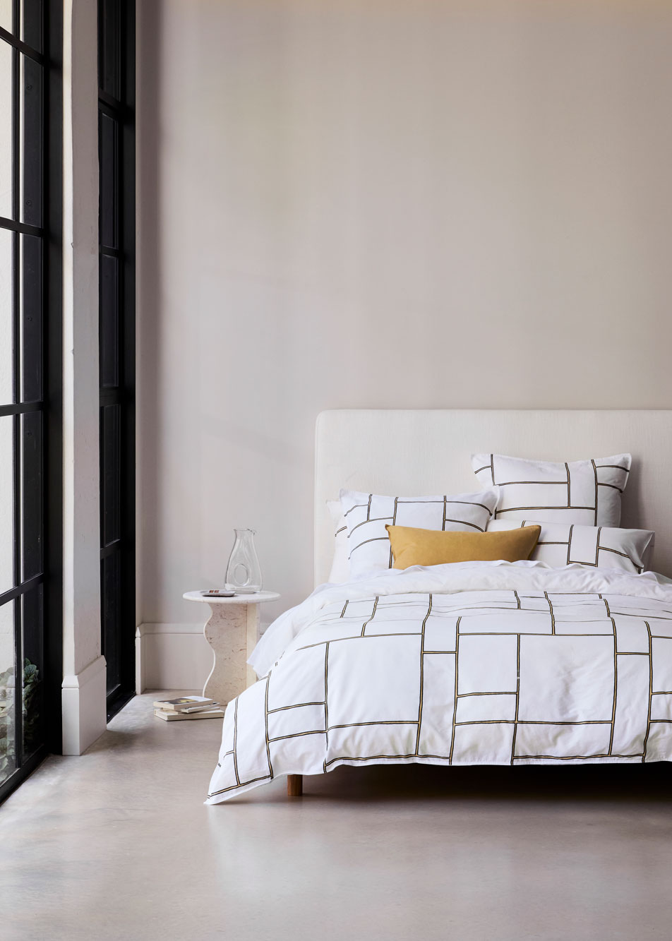 A modern bedroom with tall french windows and a concrete floor. A bed with a geometric print quilt cover and white fabric headboard sits beside a sculptural marble side table, with a glass carafe sitting on top.